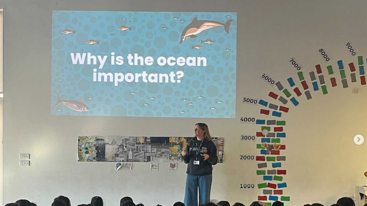A presenter stands in front of a group of children with a display above them which reads, 'why is the ocean important?'