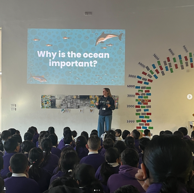 A presenter stands in front of a group of children with a display above them which reads, 'why is the ocean important?'