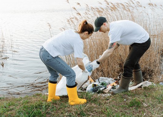 A man and woman putting litter into a white dustbin bag in front of a river. They ar wearing wellies and strong gloves.