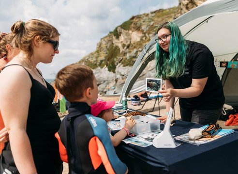 A volunteer at a Marine Conservation Society stall on a beach showing a picture of a shell to a woman and two children.