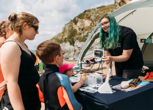 A volunteer at a Marine Conservation Society stall on a beach showing a picture of a shell to a woman and two children.