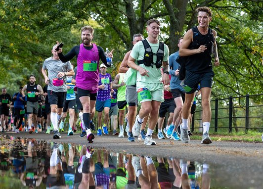 A group of runners participates in a race along a paved path lined with lush green trees. In the foreground, a large puddle reflects the vibrant colors of the runners' athletic gear.