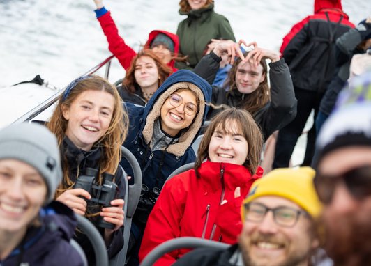 A group of smiling young people sitting in rows on a boat, one person in the foreground to the left is holding binoculars. The person in the background is making a heart with their fingers.