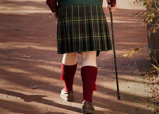 A person viewed from behind walking on a brick path, wearing a traditional Scottish kilt with a green, red, and yellow tartan pattern. They are also wearing knee-high burgundy kilt hose with tassels, brown leather shoes, and are using a dark walking cane.
