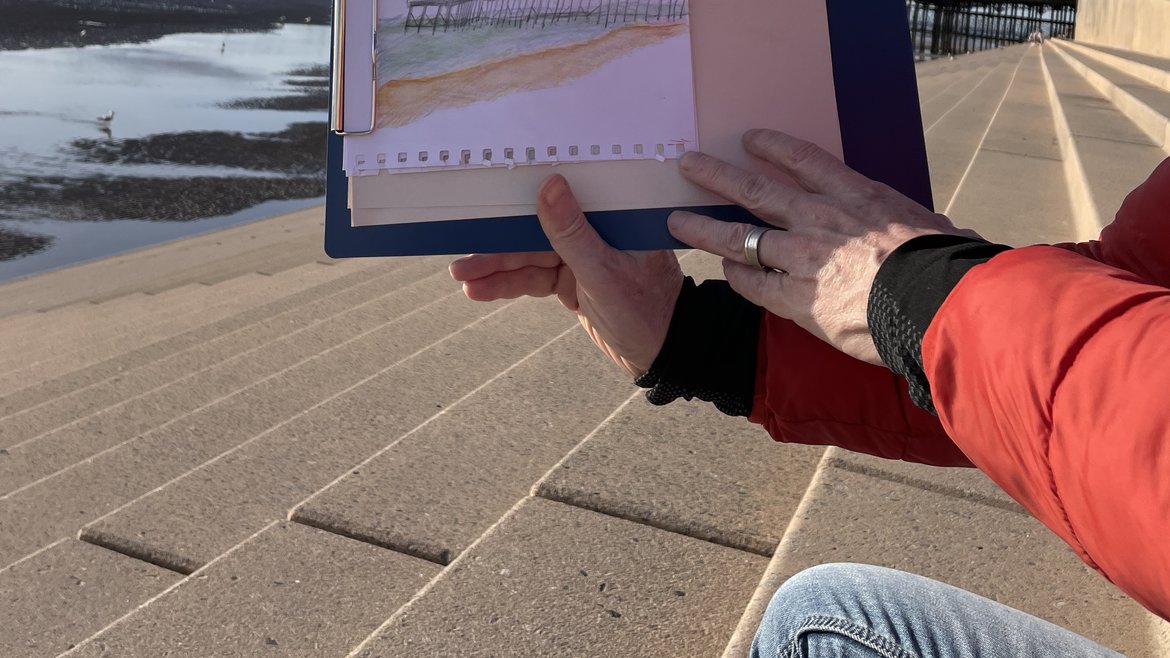 A person's hands holding up their sketch of the pier in the background to the camera