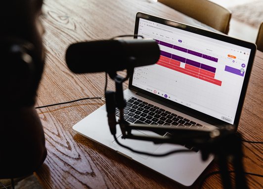 A female sits at a desk. On the desk is a laptop, and next to the laptop is a podcast microphone.