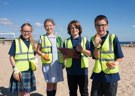 4 school children looking at the camera wearing hi-vis jackets and gloves on. The girl on the far left is holding a litter picker and the boy that is centre right is holding a clipboard. They are on a beach