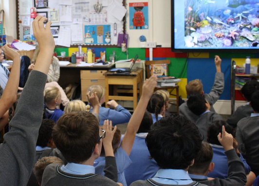 A classroom of children sitting on the floor looking at a screen, some have their hands up