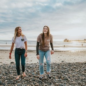 2 teenage girls laughing on the beach. It is a rocky beach with a small pier in the distance and appears to be dusk.