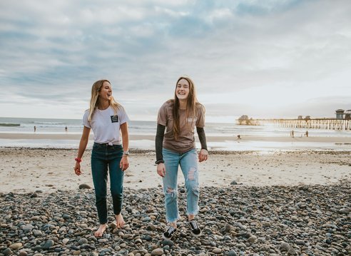 2 teenage girls laughing on the beach. It is a rocky beach with a small pier in the distance and appears to be dusk.