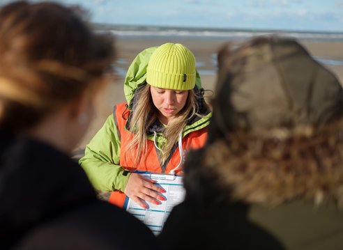 A woman wearing a lime green beanie and orange safety vest is looking down at a sheet on a clipboard. She is on a beach and is explaining the sheet to 2 people in the foreground