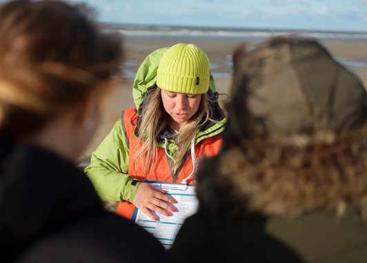 A woman wearing a lime green beanie and orange safety vest is looking down at a sheet on a clipboard. She is on a beach and is explaining the sheet to 2 people in the foreground
