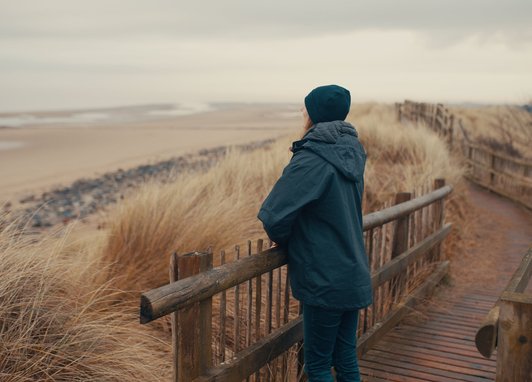 Woman standing over a fence looking towards a beach. She is wearing a dark coat and beanie. She is facing away from the photographer.