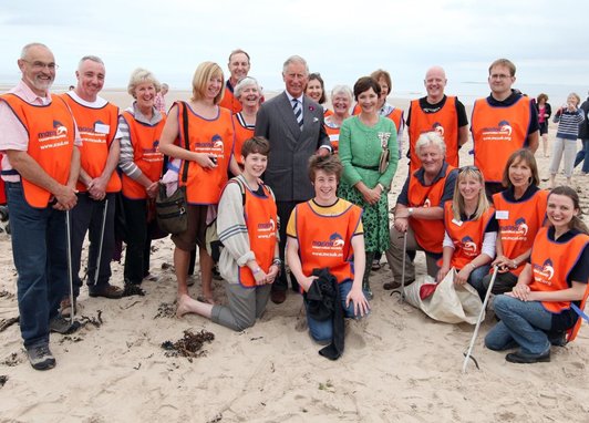 King Charles III attends a Bamburgh beach clean in 2012. He is smiling in the middle of the photo with around 20 volunteers in orange vests. Young volunteers are kneeling and sat down in the foreground.