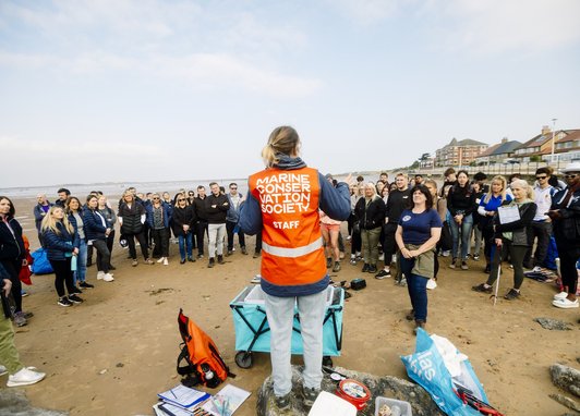 A woman wearing a hi-vis orange vest has her back to the camera. She is in front of a group of volunteers. Her vest reads Marine Conservation Society.
