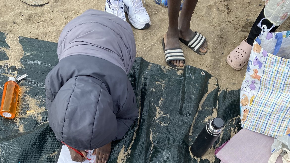 Children learning about beaches while at a beach. The shot is taken for the top down while the kids are drawing on paper. They are sitting on a dark beach towel.
