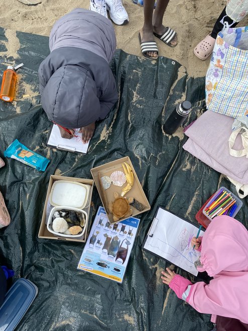 Children learning about beaches while at a beach. The shot is taken for the top down while the kids are drawing on paper. They are sitting on a dark beach towel.