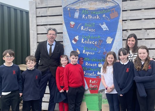 Some students and 2 adults standing in front of an outside wall with an image of a waste funnel