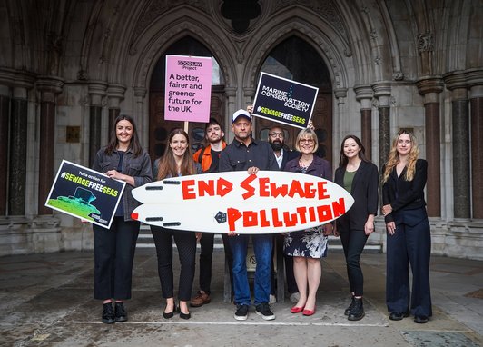 Members of the Marine Conservation Society team stand with other NGOs outside Westminster holding a surf board reading 'end sewage pollution'.