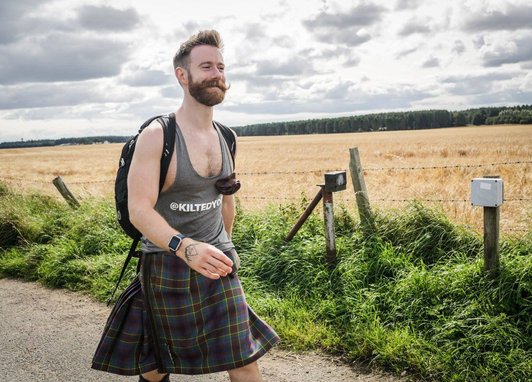 A bearded man with a handlebar mustache smiles as he walks along a rural path. He is wearing a grey tank top, a traditional tartan kilt, and a black backpack. The background features a golden field under a cloudy sky.