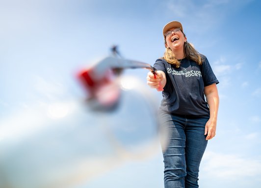 A person wearing a navy blue Marine Conservation Society t-shirt, jeans, and a tan cap laughing while using a litter picker. The camera angle is low, looking up at the person against a bright, clear blue sky. A blurred plastic bottle is in the foreground.