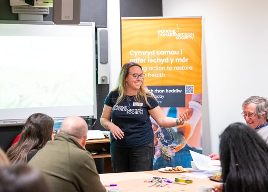 A woman wearing a Marine Conservation Society t-shirt speaking to an adult audience in front of a banner that reads 'Cymryd camau i adfer iechyd y môr' and 'taking action to restore marine health'."