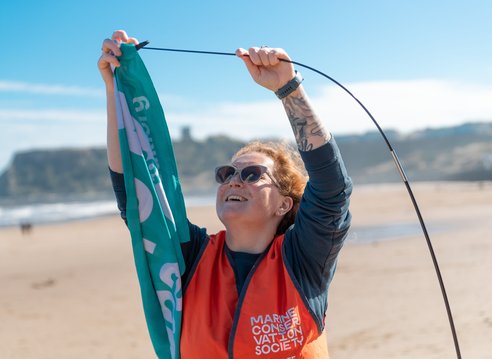 A woman wearing snglasses and an orange hi-vis vest is placing a teal flag onto a pole. Behind he is a beach.