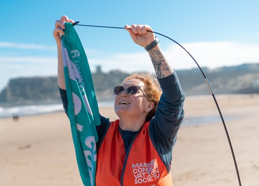 A woman wearing snglasses and an orange hi-vis vest is placing a teal flag onto a pole. Behind he is a beach.