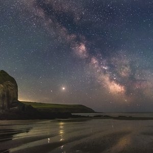The night sky is shining with stars and the moon. Mothecombe coastline can be seen to the left of the photo, and the watery sand is reflecting the dark sky.