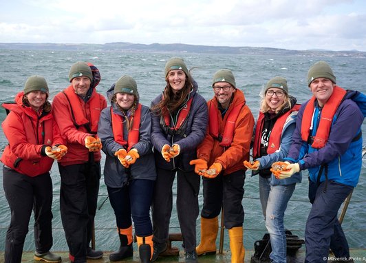 Seven scientists and Marine Conservation Society staff involved in the Restoration Forth project stand smiling on the Firth of Forth shore. They are all wearing protective orange gloves with European oysters in their spread-out palms.