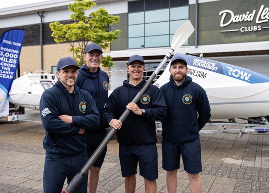 4 men standing in front of a boat. The centre right man is holding a boat paddle. They are all wearing caps and 'Give us a wave' branded hoodies.