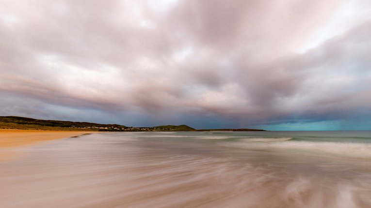 An expansive, long-exposure photograph of Narin Strand, a sandy beach, under a dramatic, overcast sky. The motion blur of the receding tide creates smooth, misty white streaks across the foreground.