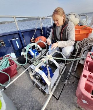 A Marine Conservation Society scientist sets up a BRUV device from a boat deck. The device is mechanical and looks like a motor surrounded by a cage.