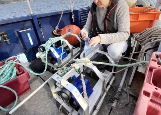A Marine Conservation Society scientist sets up a BRUV device from a boat deck. The device is mechanical and looks like a motor surrounded by a cage.