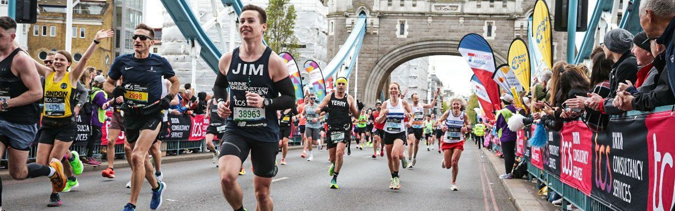 Jim Bourne wears a dark blue marine conservation society running vest. He is photographed running the London Marathon. Tower bridge can be seen behind him along with lots of other runners