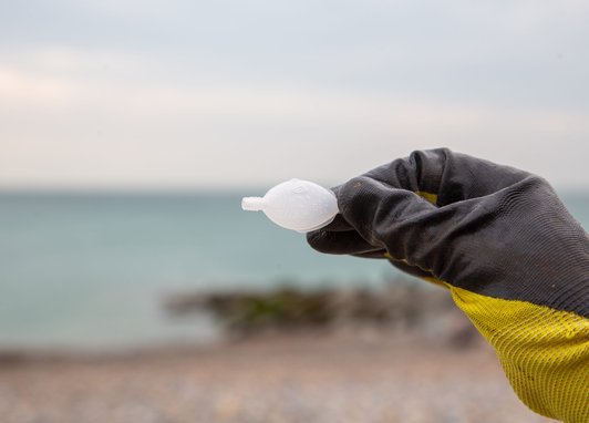 A gloved yellow hand holding an empty single-use plastic fish-shaped soy sauce container. In the background is a beach