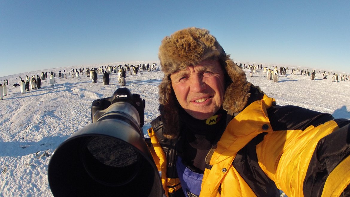 Doug Allen smiles at the camera and points his own huge camera lens at the photographer. He is at an emperor penguin colony and a hundred plus penguins can be seen behind him. Naturally it's cold, with ice and a bright blue sky!