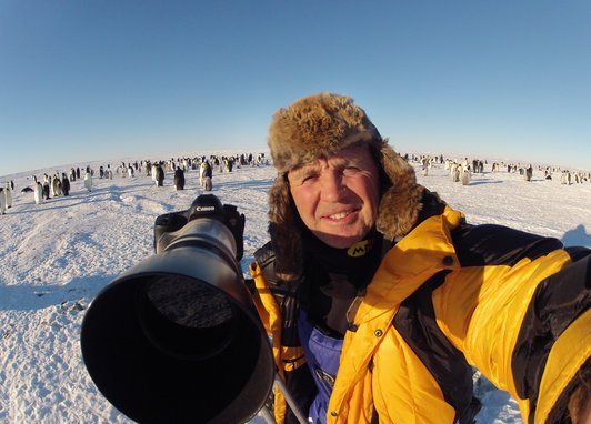 Doug Allen smiles at the camera and points his own huge camera lens at the photographer. He is at an emperor penguin colony and a hundred plus penguins can be seen behind him. Naturally it's cold, with ice and a bright blue sky!