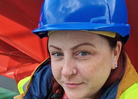 A picture of Bev wearing a hard hat and hi-vis jacket looking into the camera