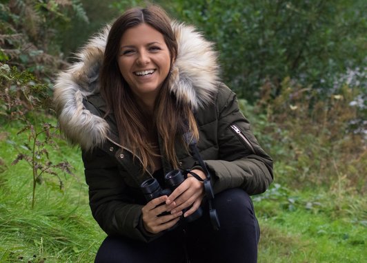 Lizzie Daly smiles at the camera against a green grass and woodland setting. She is wearing a puffer jacket with a fur hood.