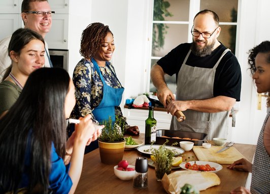 A group of adults are standing around a kitchen island, laughing and chatting as a man holding a pepper grinder puts the finishing touches to a tasty seafood meal.