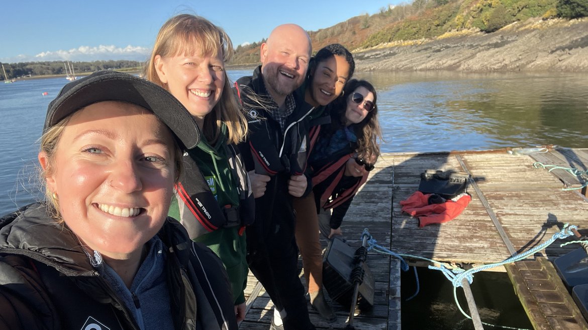 4 staff members from the Natur Am Byth porject stand huddled together on a jetty, smiling at the camera, as the sun shines behind them