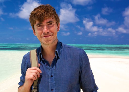 Simon Reeve stands smiling in a blue shirt against a backdrop of a white sand beach and clear blue ocean.