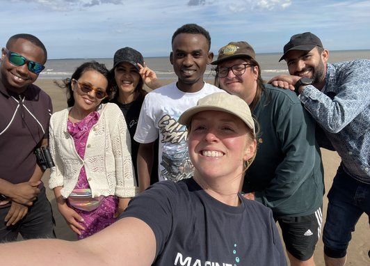 A selfie taken on a camera with 7 people posing for the camera. Behind them is a sunny beach. The girl taking hte picture is wearing a Marine Conservation Society t-shirt.