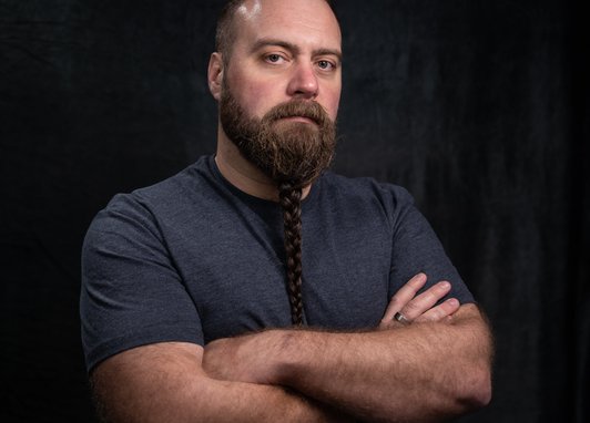 Tom 'The Blowfish' Hird looks at the camera without smiling. He has his arms crossed against a dark backdrop. His long beard is braided and his muscular arms can be seen as he is wearing a grey tshirt.