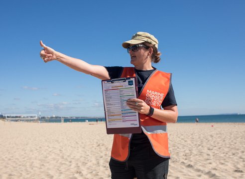 A woman with a hat and sunglasses standing on a beach. She is pointing to the left while holding a survey form for others to see. She is wearing a bright orange safety vest with a Marine Conservation society logo on it.