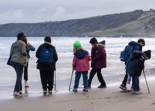Children wearing coats and backpacks walking along the coast taking part in a beach clean. Some are holding litter pickers. Some cliffs are visible behind them.