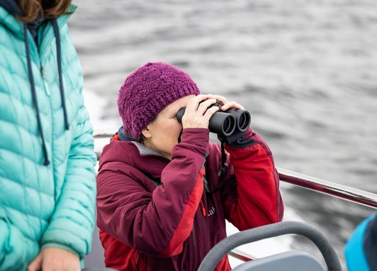 A person in a red jacket and purple beanie is using black binoculars, with another person in a teal jacket partially visible nearby. They are on a boat.