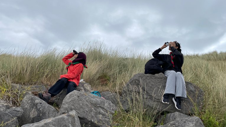2 people sititng on a grassy hill looking upward through binoculars. There are a few big stones in front of them on the hill.
