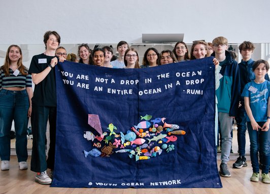 A group of young adults holding up a big tapestry with fish on it. At the top it reads "You are not a dop in the ocean you are an entire ocean in a drop - Rumi". At the bottom it reads "#Youth Ocean Network"